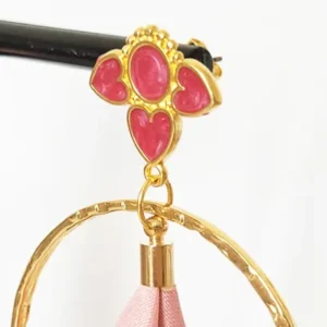 Boucles d'oreilles pendantes avec des supports en formes de 3 coeurs colorés en prune nacrée et dorés. Ces supports ont un anneau martelé et un pompon en imitation satin de couleur rose à l'intérieur.