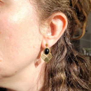 Vue portée des boucles d'oreilles dorées pendantes avec cabochon ovale noir en verre et triangle doré ajouré