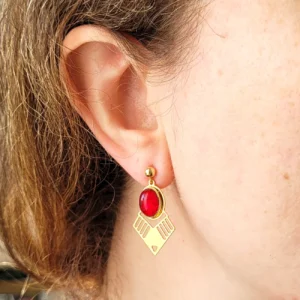 Vue portée des boucles d'oreilles dorées pendantes avec cabochon ovale rouge en verre et triangle doré ajouré